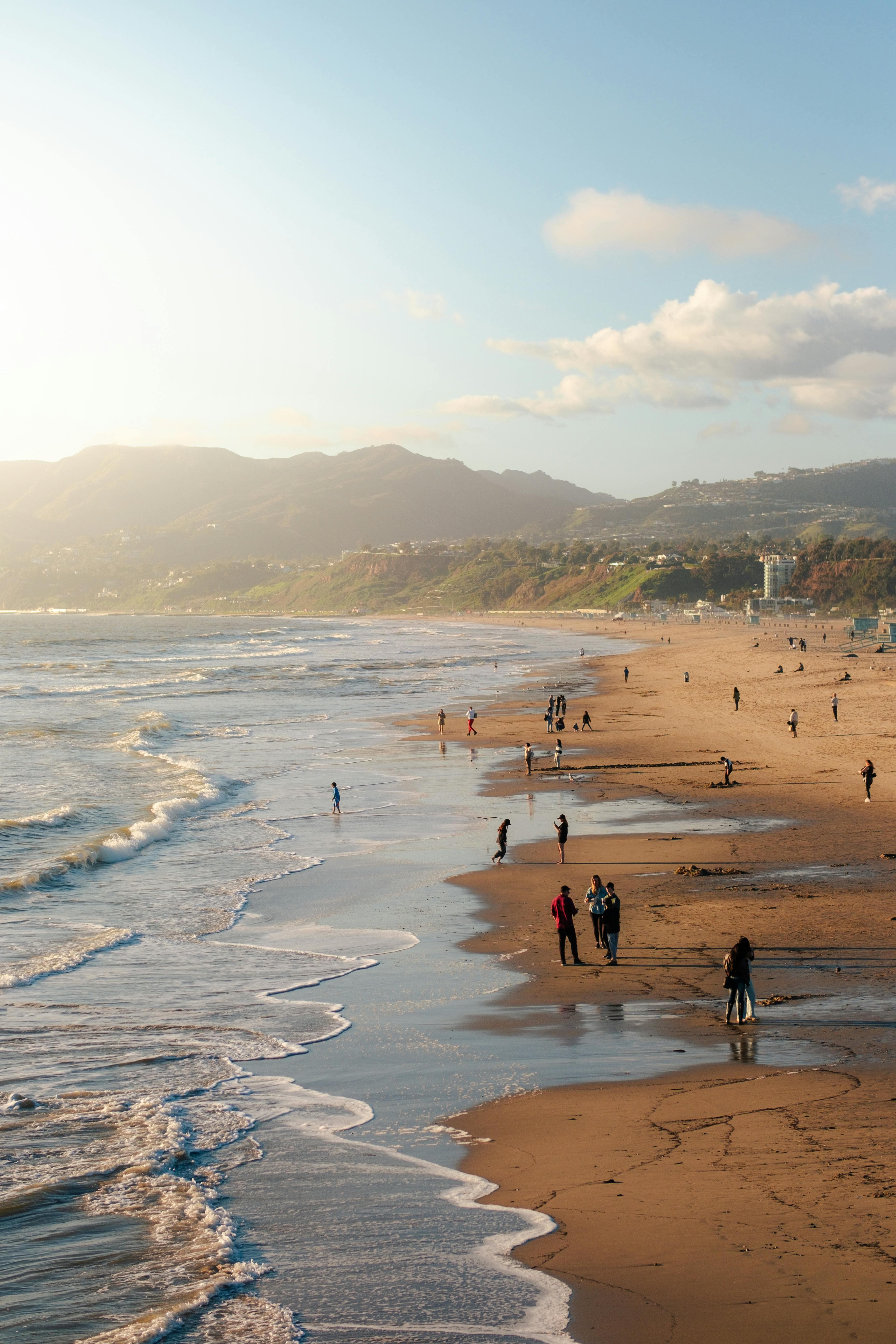 People walking along a sandy beach at sunset with waves and mountains in the background